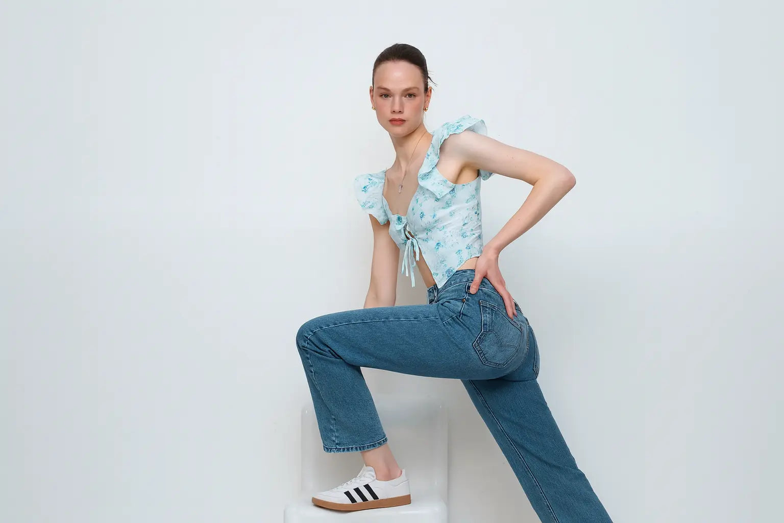 Woman wearing a light blue top and jeans sitting on a white box against a white background.